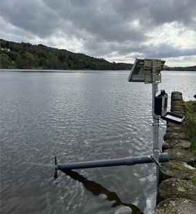 In the foreground, a Proteus telemetry system peli case and solar panel installed on a pole and a stilling tube installed in the lake. 